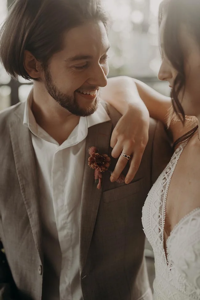 Couple de mariés complices dans des tons neutres et lumineux, l'homme portant un costume beige et une boutonnière fleurie.