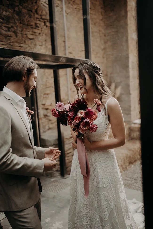 Un couple de mariés souriants s'échange un regard complice, la mariée tenant un bouquet coloré devant un mur de pierre.