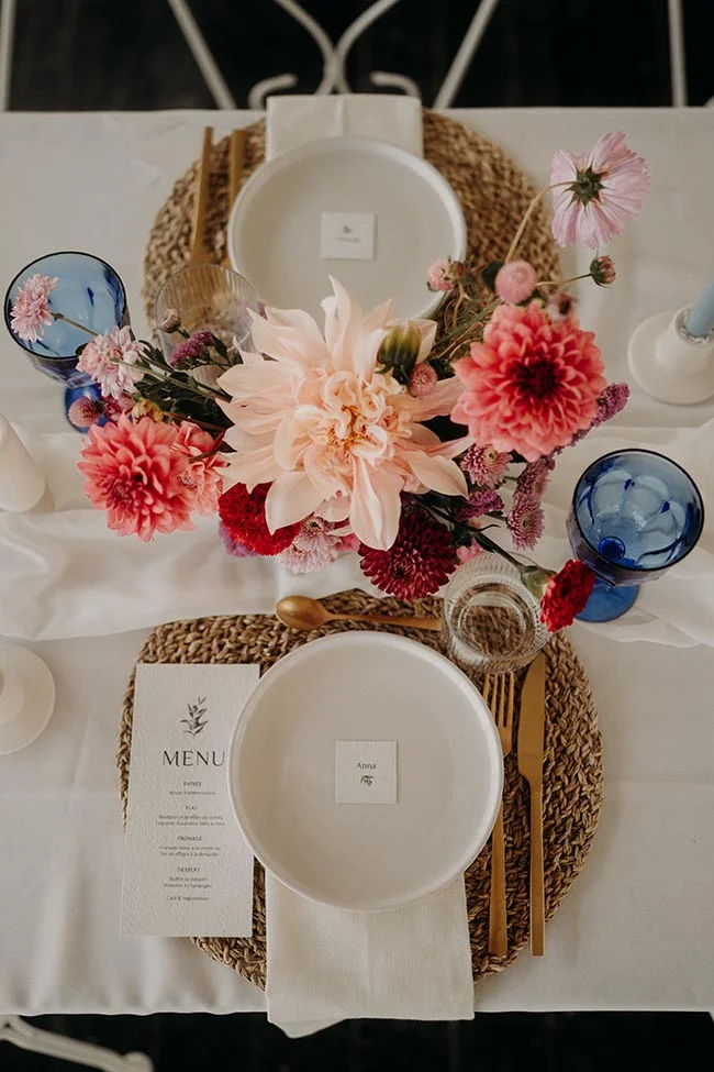 Table dressée avec des fleurs roses, de la vaisselle blanche et bleue et des sets de table en osier, ambiance estivale.