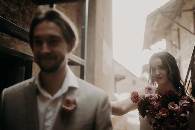 Un couple de mariés souriant dans un décor rustique, avec un bouquet de fleurs colorées aux tons automnaux.