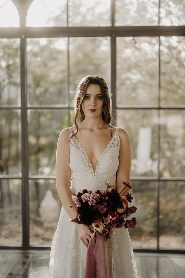 Mariée au regard intense en robe blanche dentelée, tenant un bouquet sombre devant une grande verrière lumineuse.