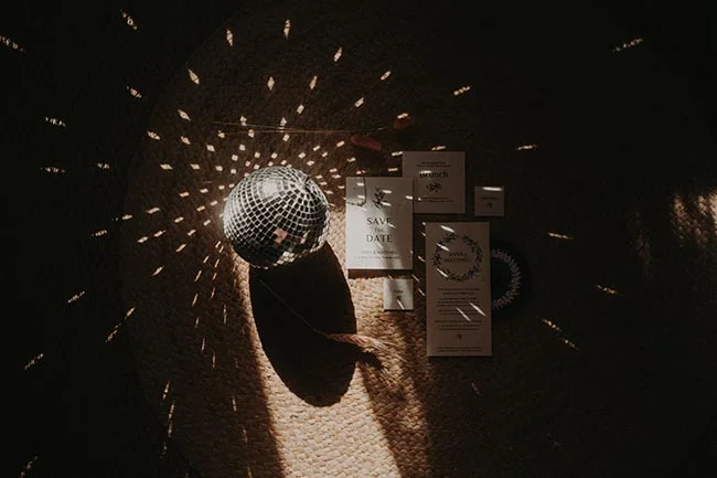 Boule à facettes et faire-part de mariage sur un fond texturé, éclairés par des jeux de lumière dorée et tamisée.