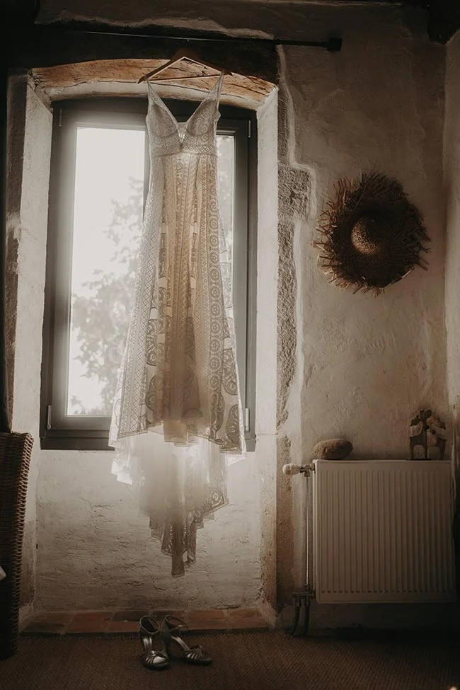 Robe de mariée en dentelle blanche suspendue devant une fenêtre dans une pièce rustique à l'ambiance bohème.