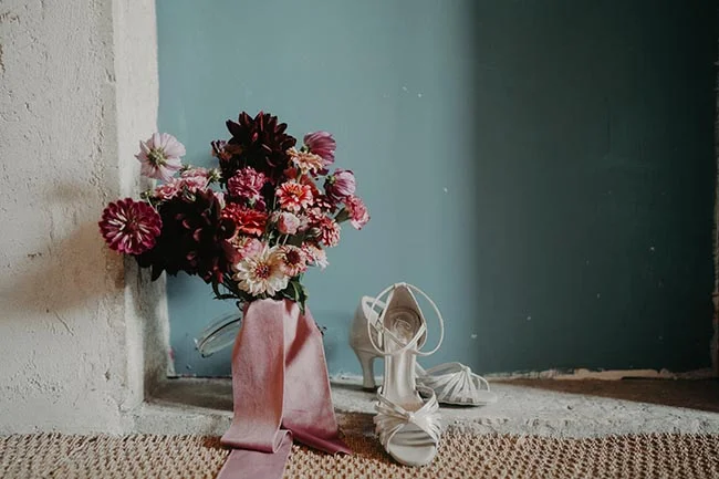 Bouquet de fleurs roses et bordeaux et sandales blanches à talons sur un fond bleu pastel, ambiance romantique.