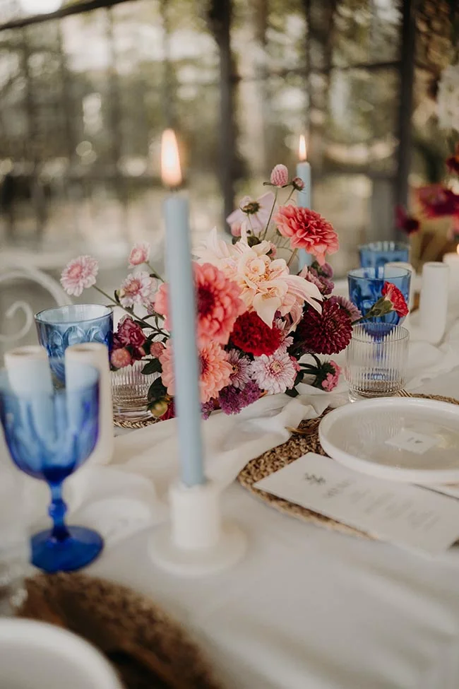 Table élégante avec fleurs roses et rouges, bougies bleues et verres assortis pour un mariage aux tons méditerranéens.