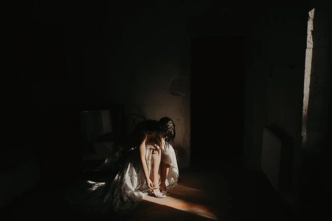 Une mariée en robe blanche s'habille dans une pièce sombre, éclairée par un rayon de lumière dramatique.