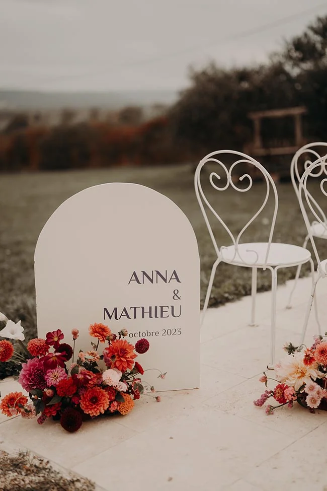 Panneau de bienvenue blanc pour Anna et Mathieu avec fleurs colorées et chaises en fer forgé dans un jardin.