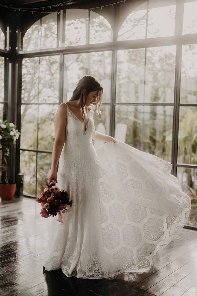 Une mariée en robe blanche dentelée fait tournoyer sa jupe devant une grande baie vitrée, tenant un bouquet automnal.