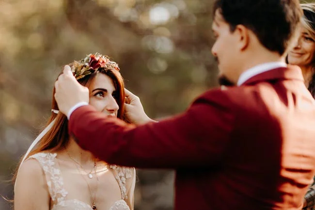 Un marié en costume bordeaux place une couronne de fleurs sur la tête de sa future épouse lors d'un elopement romantique.