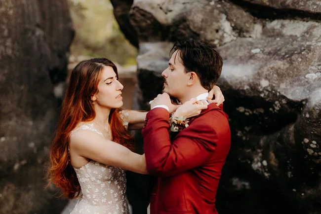 Couple d'élopement s'enlaçant tendrement devant des rochers, elle en robe blanche et lui en costume rouge bordeaux.