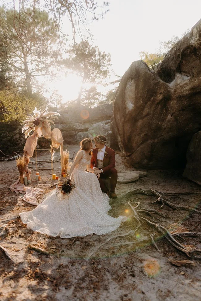 Couple de mariés en tenue élégante posant dans un décor bohème et rocheux sous une lumière dorée et naturelle.