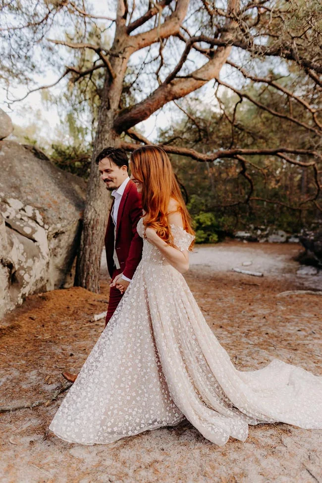 Couple marié s'enlaçant dans une forêt, elle en robe blanche scintillante et lui en costume bordeaux.