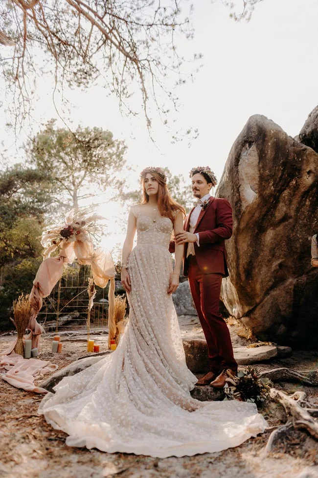 Couple de mariés en tenue bohème posant devant un arche fleurie et des rochers dans une ambiance naturelle et romantique.