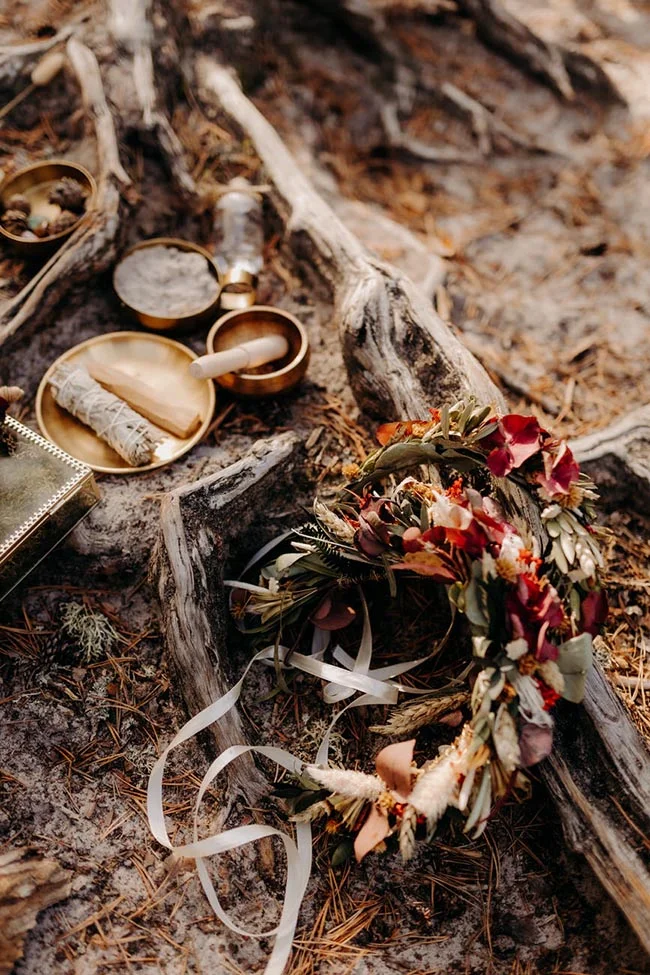 Couronne de fleurs séchées et accessoires rituels dorés posés sur un vieux tronc d'arbre dans une ambiance naturelle.