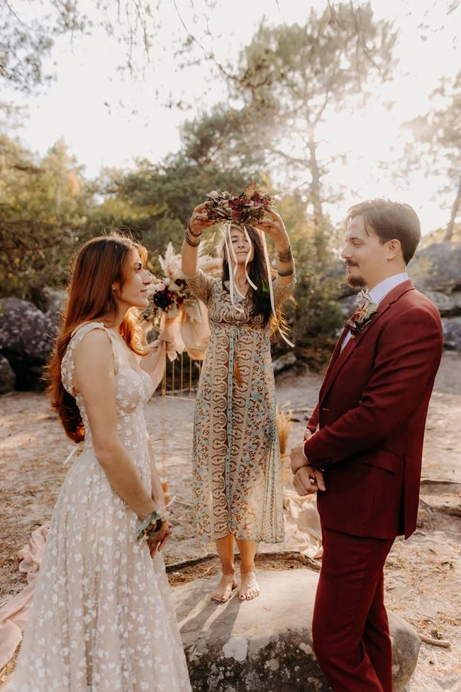 Couple en tenue de mariage et officiante sous une couronne de fleurs dans un cadre naturel et lumineux.