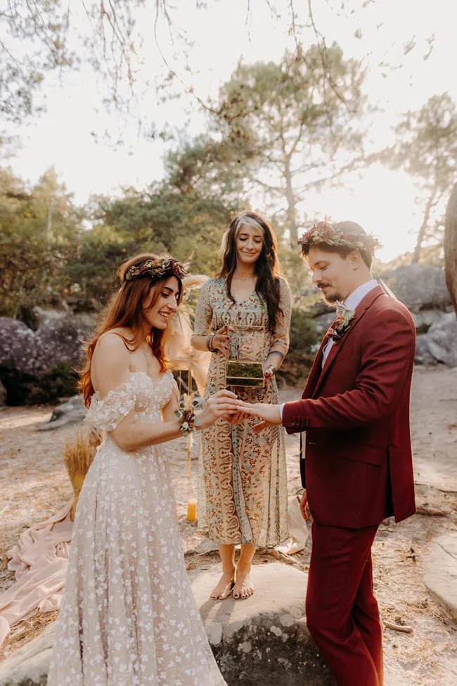 Couple en tenue de mariage et officiante échangeant des alliances en forêt dans une ambiance bohème et romantique.