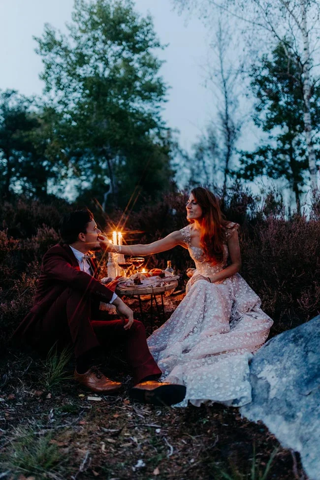 Couple en tenue de mariage partageant un dîner romantique aux chandelles en pleine nature au crépuscule.