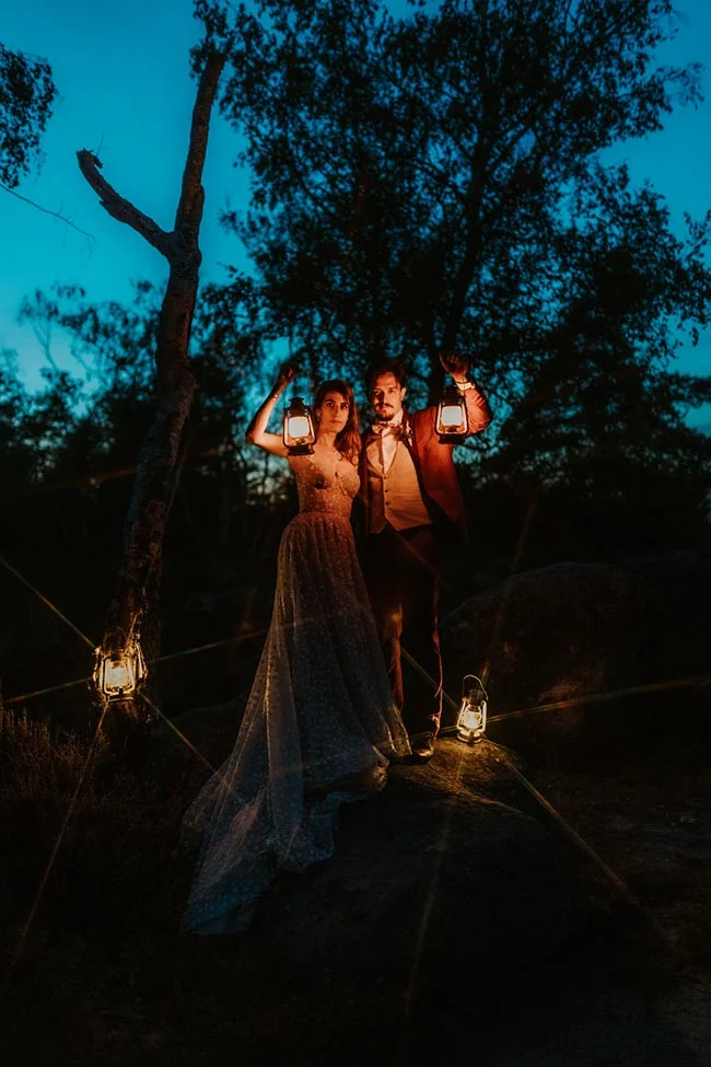 Couple en tenue de mariage tenant des lanternes dans une forêt sombre sous un ciel bleu crépusculaire.