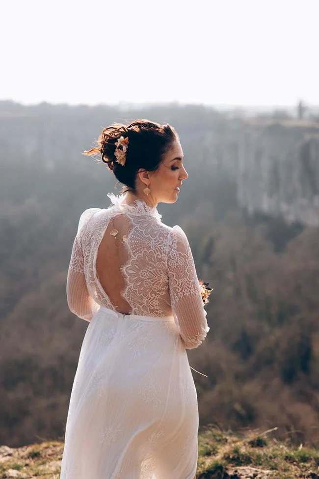 Mariée de profil en robe blanche en dentelle, cheveux relevés avec fleurs, face à un paysage naturel brumeux.