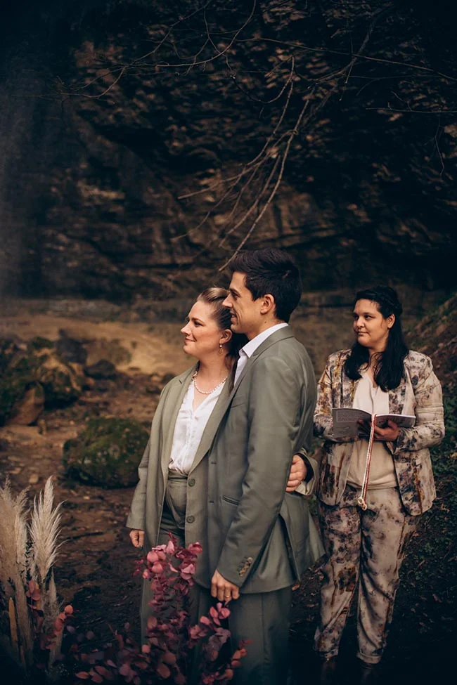 Couple en costumes vert sauge et officiante dans un décor naturel sombre et rocheux.