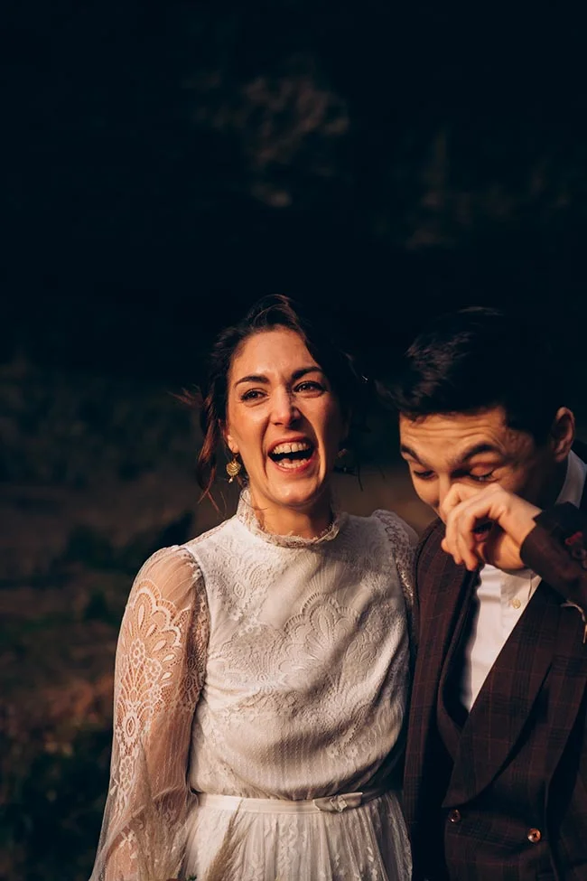 Jeune couple riant avec complicité, la mariée en robe de dentelle blanche et le marié en costume sombre.