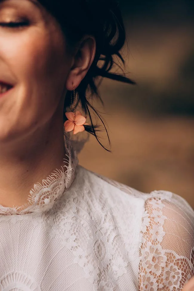 Gros plan d'une mariée en robe de dentelle blanche portant des boucles d'oreilles fleurs pêche.