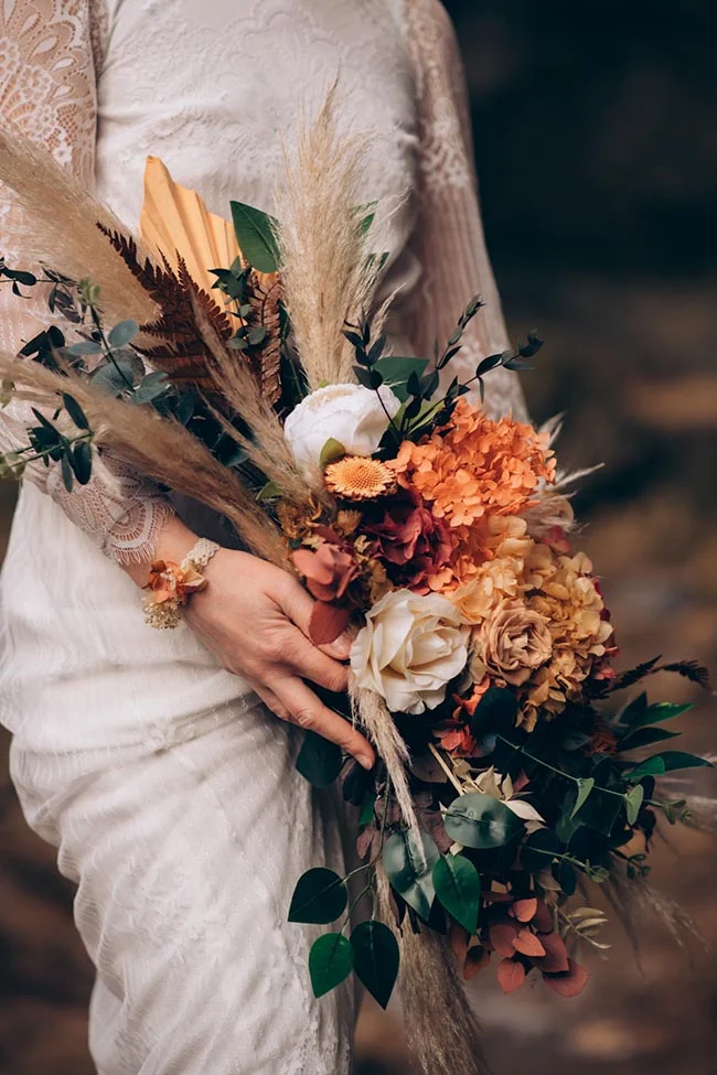 Mariée en robe blanche tenant un bouquet bohème aux tons terracotta, crème et eucalyptus.