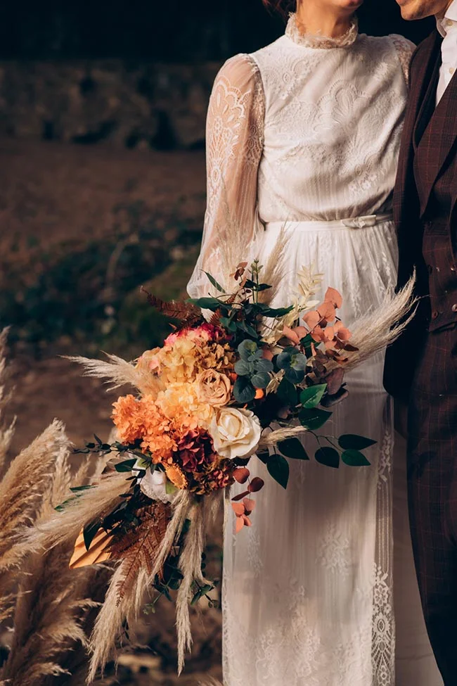 Couple en tenue de mariage tenant un bouquet bohème aux tons orangés et crème dans un décor naturel.