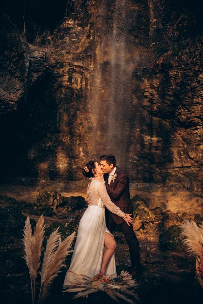 Couple de mariés s'embrassant devant une cascade illuminée dans un décor rocheux et romantique.