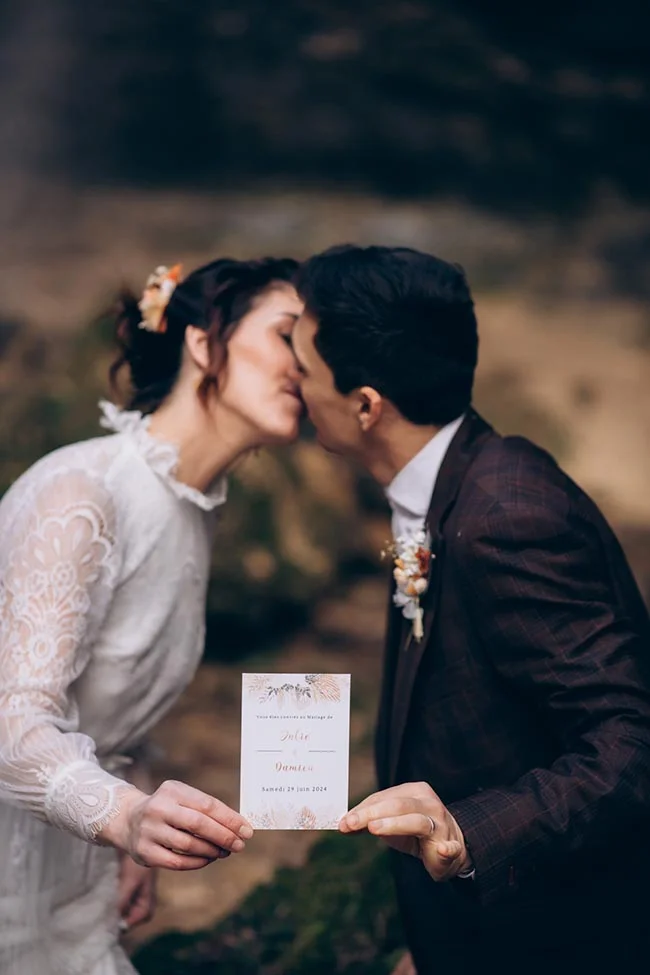 Jeunes mariés s'embrassant tendrement tout en tenant leur faire-part, dans une ambiance romantique et naturelle.