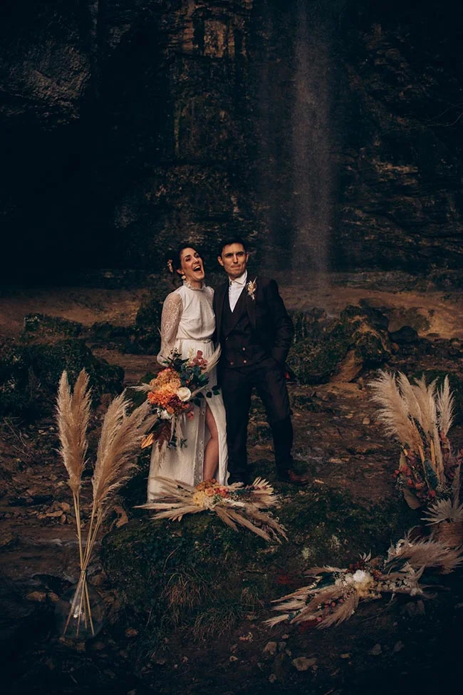 Couple marié riant devant une cascade sombre, entouré de fleurs séchées et de pampas dans une ambiance naturelle et intime.