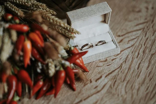 Deux alliances dans un écrin blanc posées sur du bois, à côté d'un bouquet de piments rouges et d'épis de blé.