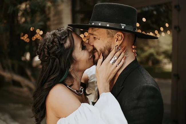 Couple complice s'embrassant tendrement, lui en costume et chapeau noir, elle en robe blanche, ambiance romantique.