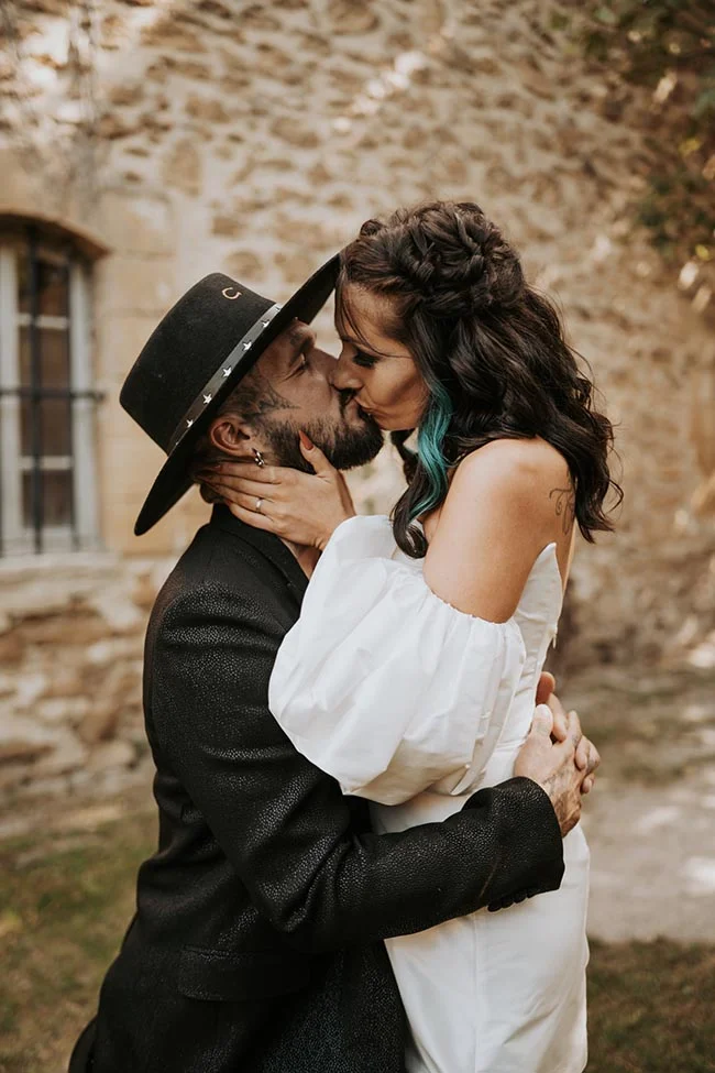 Couple glamour s'embrassant avec passion devant un mur de pierre, lui en costume noir et chapeau, elle en robe blanche.