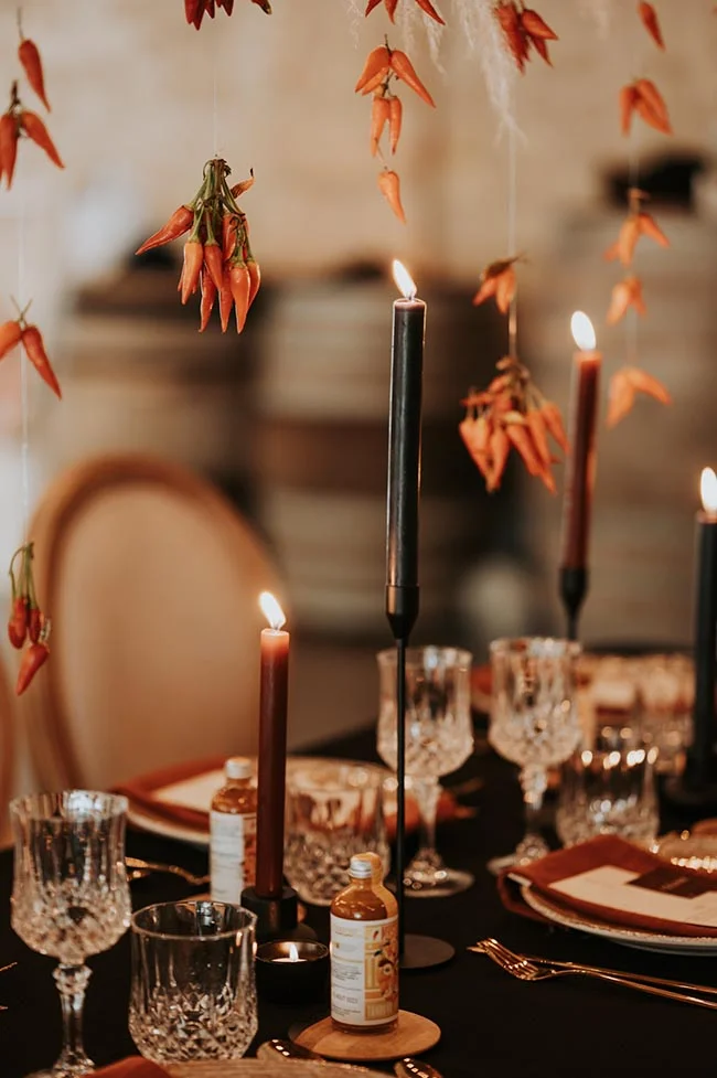 Décoration de table sombre et élégante avec bougies fines, verres en cristal et piments rouges suspendus.