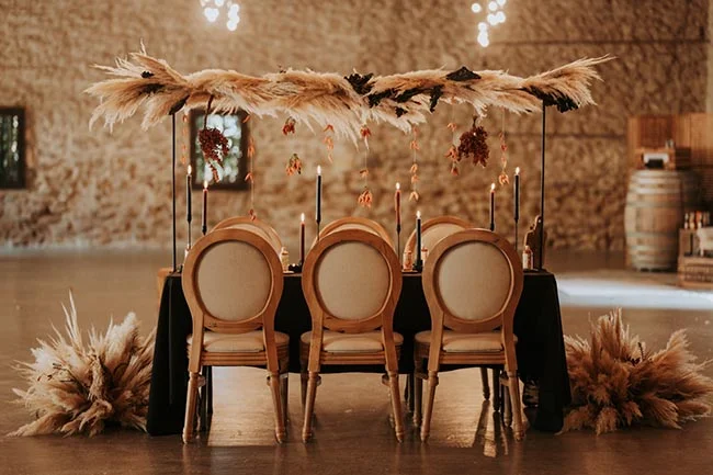 Table de mariage élégante avec chaises beiges, bougies et arche de pampas dans un cadre rustique et chaleureux.