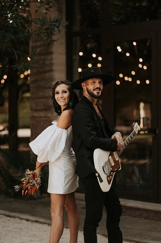 Couple marié au style rock : elle en robe blanche courte, lui en costume noir avec chapeau et guitare électrique.