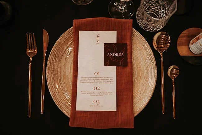 Dressage de table élégant avec menu, couverts cuivrés et serviette terracotta sur fond noir pour un mariage spicy.