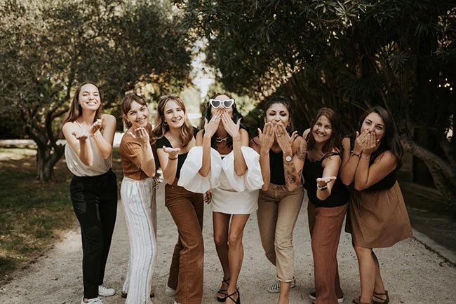 Six jeunes femmes souriantes et élégantes en tenues neutres lancent des baisers dans un jardin ensoleillé.