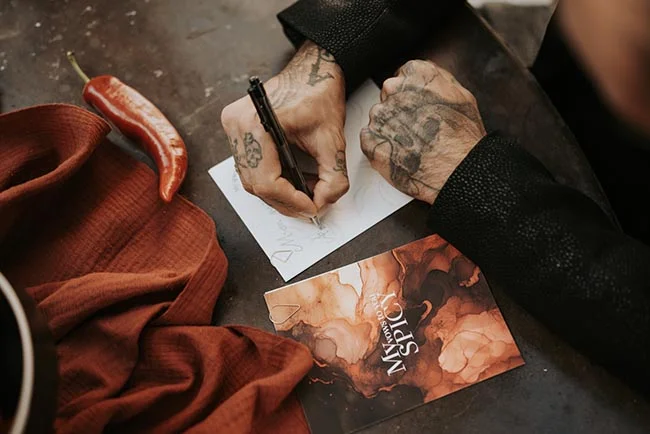 Gros plan sur des mains tatouées écrivant sur un carton d'invitation blanc à côté d'un piment rouge et d'un tissu terracotta.