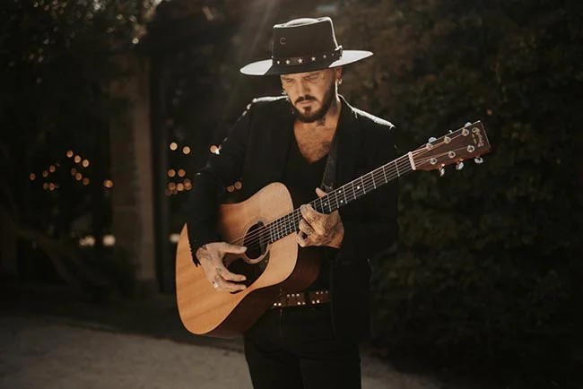 Homme élégant en costume noir et chapeau jouant de la guitare acoustique dans une ambiance nocturne et tamisée.