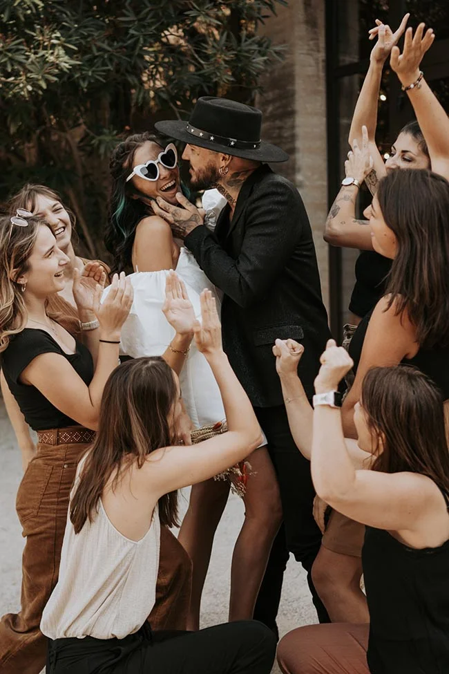 Un couple excentrique s'embrasse entouré d'amis lors d'un mariage festif et moderne aux tons neutres et noirs.