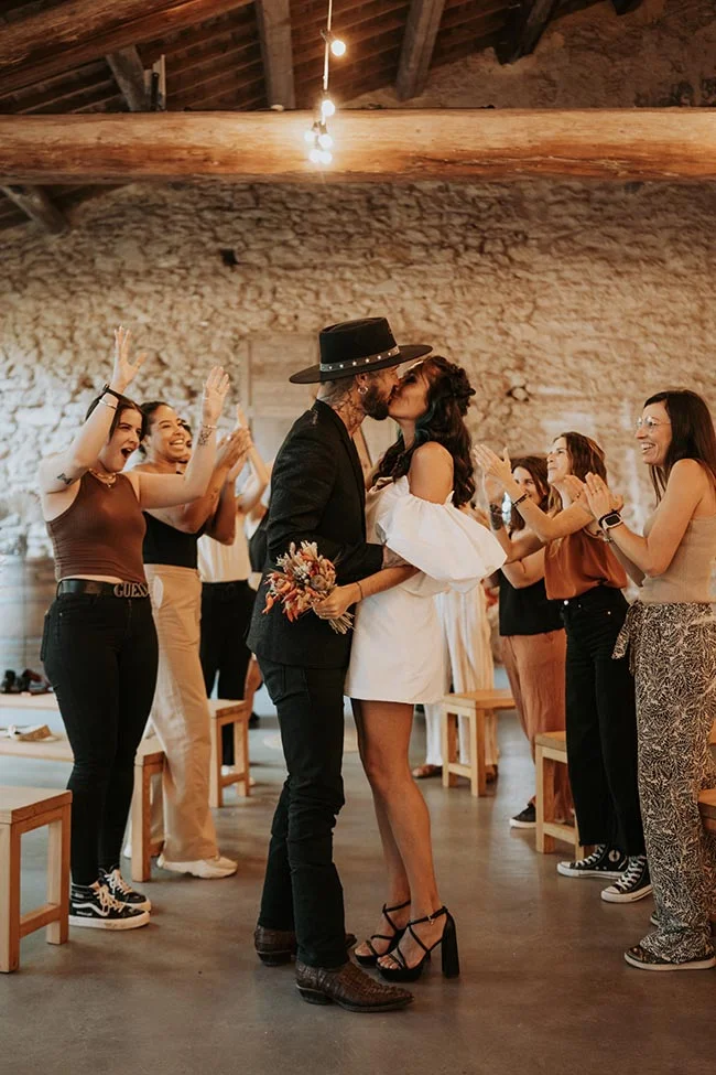 Un couple s'embrasse avec passion dans une grange rustique, entouré d'invités enthousiastes et applaudissants.