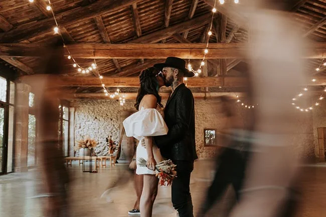 Couple s'embrassant dans une grange rustique, elle en robe blanche courte et lui en costume noir avec chapeau.