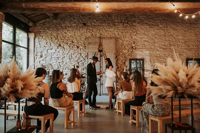 Couple échangeant des vœux dans une salle en pierre rustique, entouré d'invités et de pampas beiges.