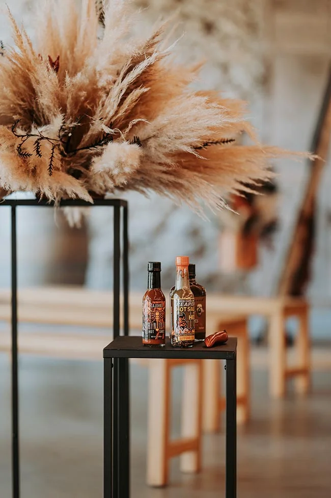 Trois bouteilles de sauce piquante sur un support noir, sous un bouquet de pampas beiges dans un décor chaleureux.