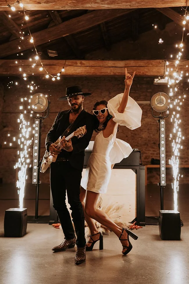 Couple marié excentrique et joyeux posant avec une guitare électrique sous des étincelles dans un cadre rustique.