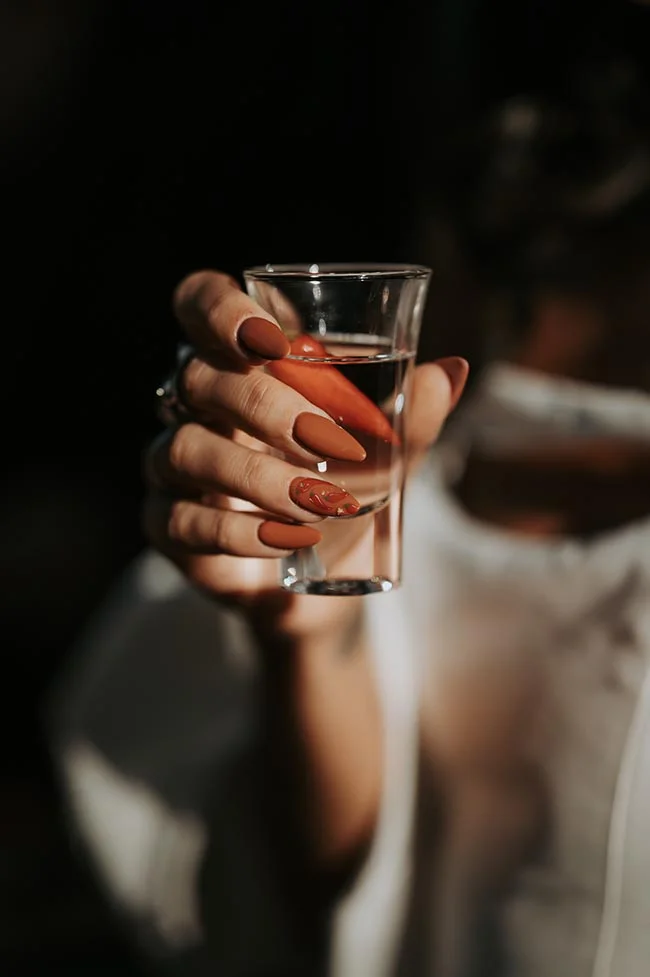 Main aux ongles terracotta tenant un verre de tequila avec un piment rouge, ambiance chaleureuse et festive.
