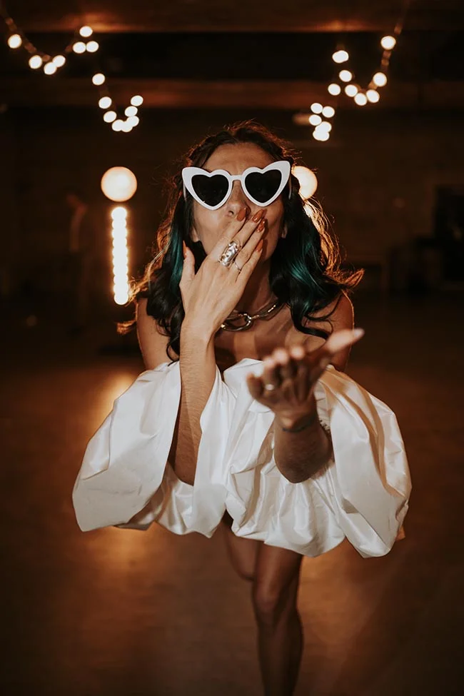 Femme en robe blanche et lunettes cœur, posant avec malice dans une ambiance festive et tamisée.