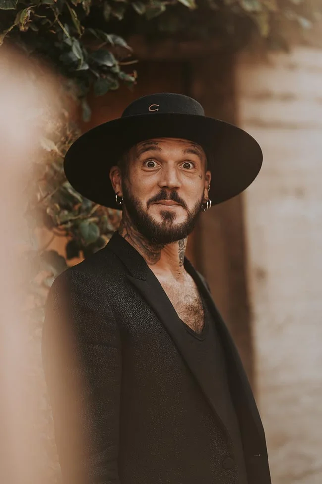 Homme au look audacieux en costume noir et chapeau à larges bords, arborant des tatouages et un regard expressif.